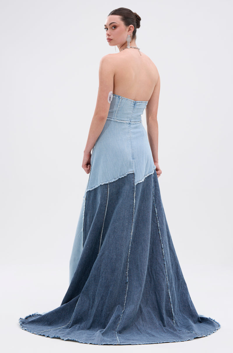 A woman in the IT'S BRITNEY PATCHWORK DENIM MAXI DRESS, featuring a strapless fitted bodice and flared mermaid-style floor-length skirt in varying blue denim shades, stands facing away and glances over her shoulder.