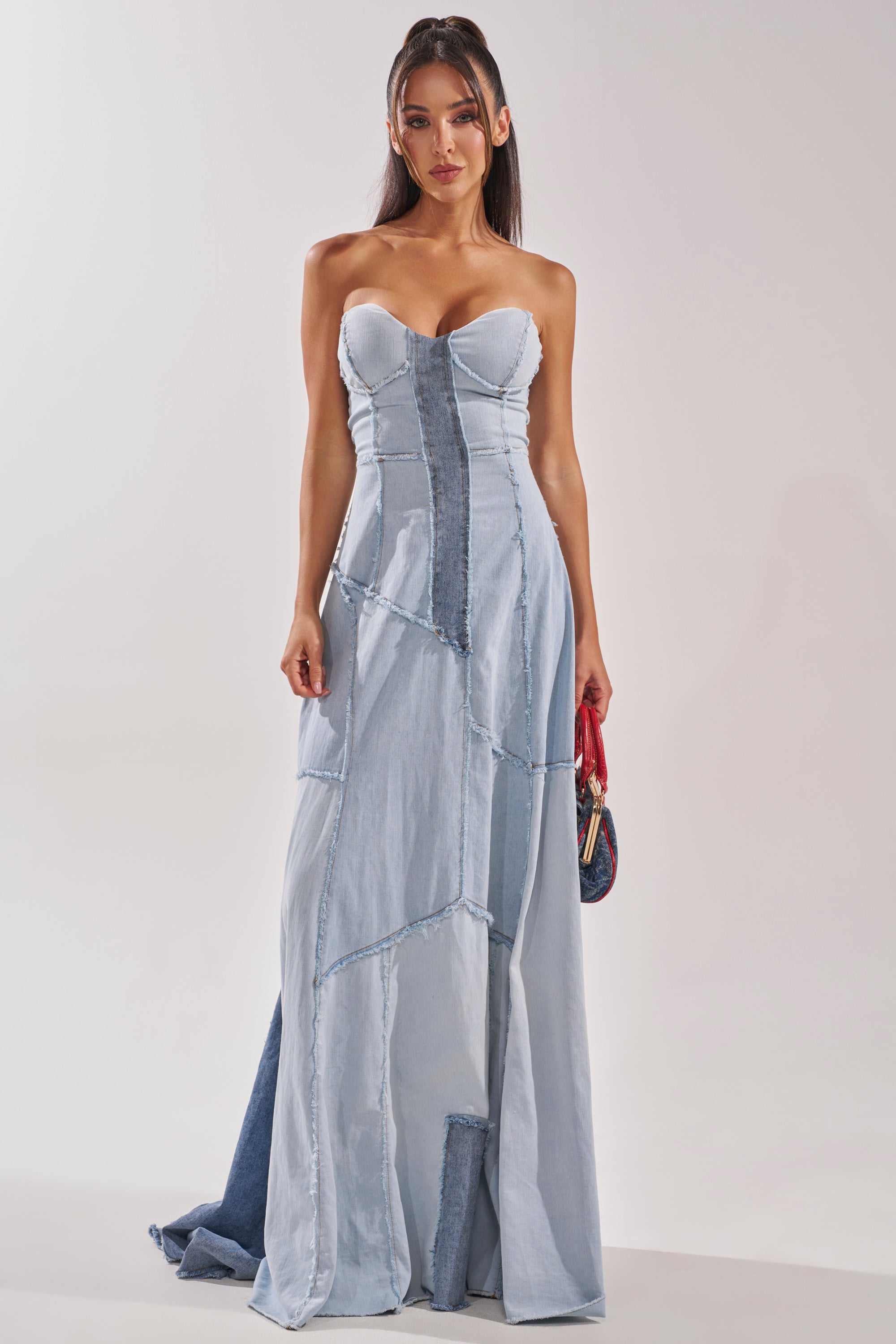 A woman wears the IT'S BRITNEY PATCHWORK DENIM MAXI DRESS, featuring a strapless fitted bodice and flared skirt. She holds a small red purse, her long dark hair in loose waves, against a plain light background. 3