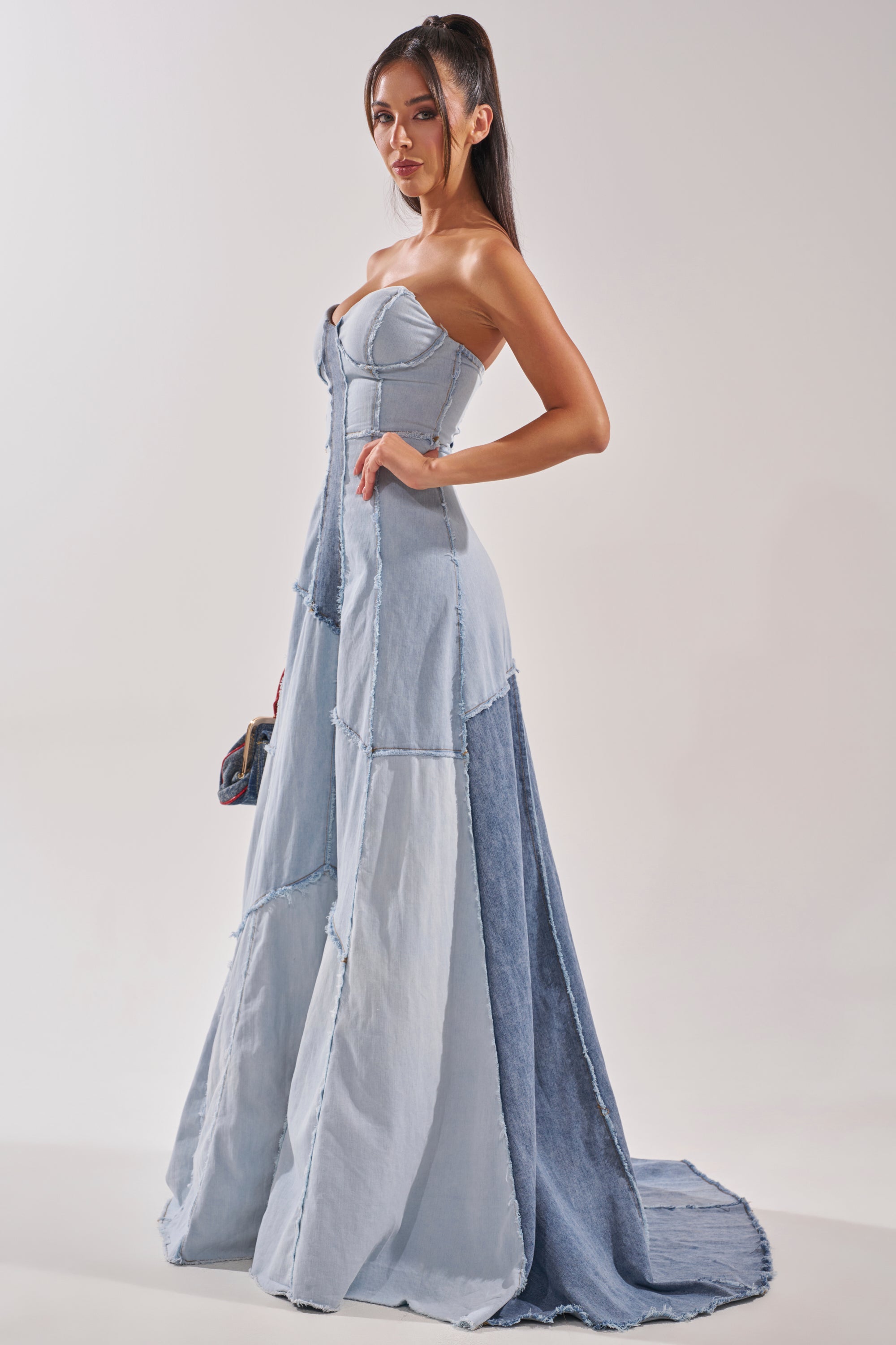 A woman with long, dark hair in a high ponytail stands holding a small clutch, wearing the IT'S BRITNEY PATCHWORK DENIM MAXI DRESS—a light blue, strapless piece with a fitted bodice and flared, floor-length skirt for Y2K popstar vibes. 4