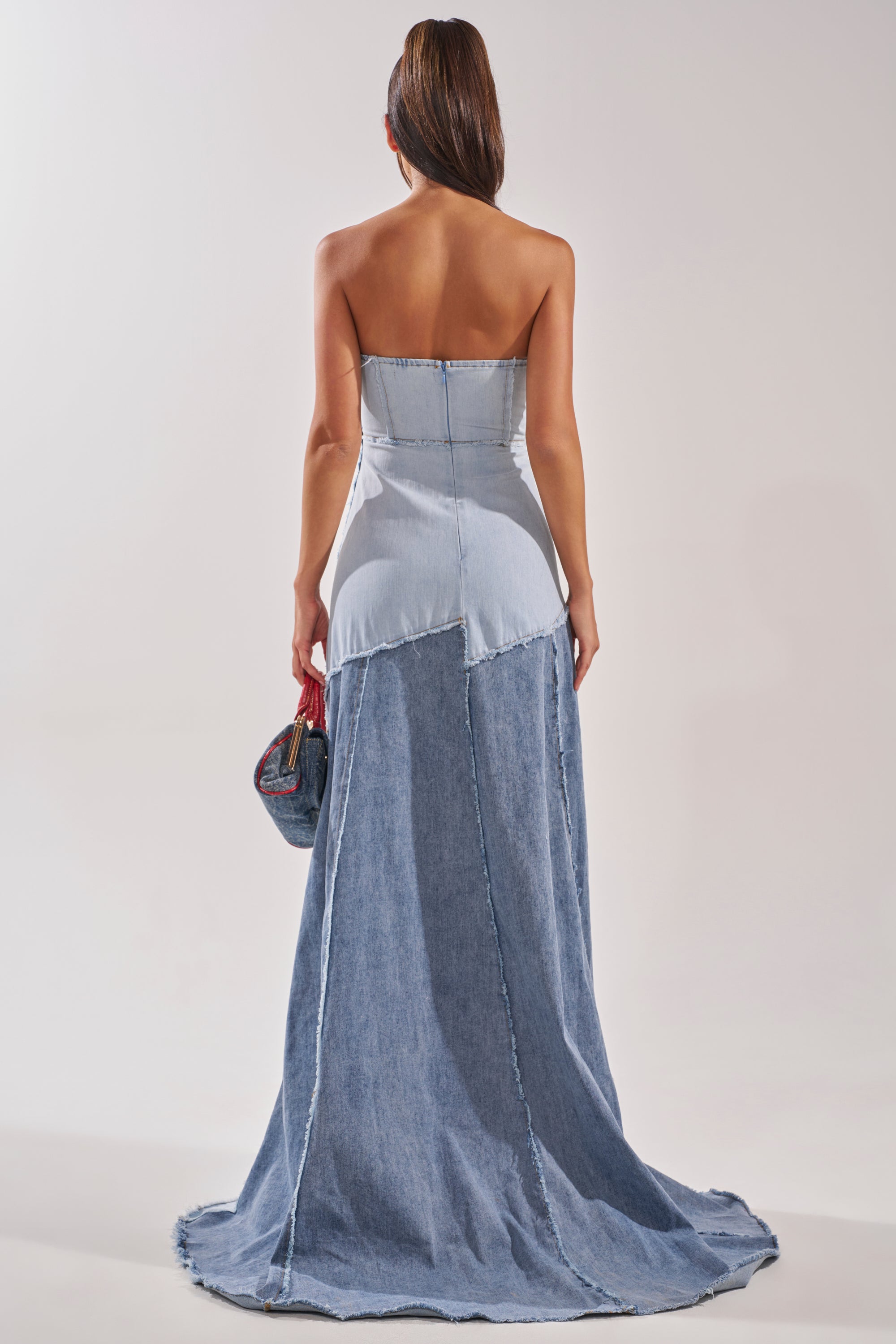 A person with long hair in a ponytail stands facing away, wearing the IT'S BRITNEY PATCHWORK DENIM MAXI DRESS—a strapless, floor-length denim look—while channeling Y2K popstar vibes and holding a small handbag in their right hand. 5