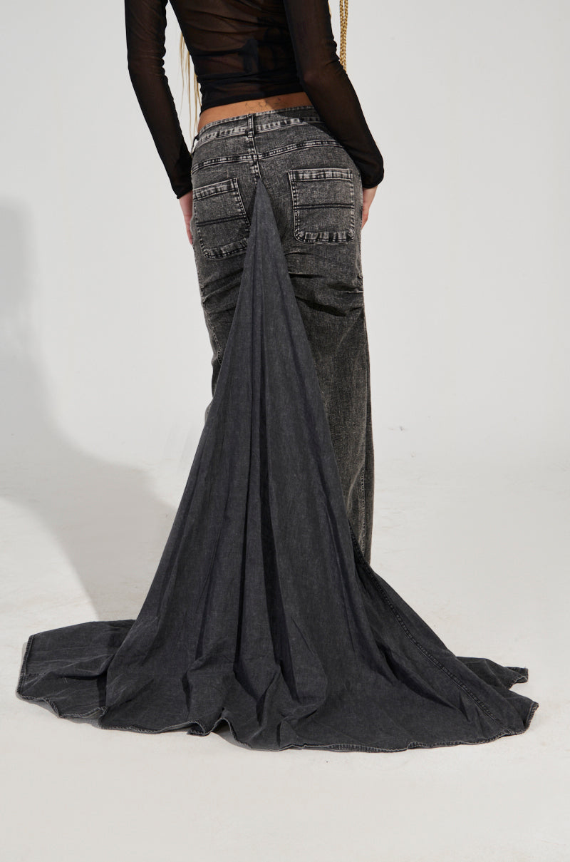 A person is wearing the "OH THE DRAMA DENIM MAXI SKIRT WITH TRAIN IN GREY," featuring a distinctive long, triangular train. Paired with a sheer black top, the skirt's high-rise waist and ruched back detailing elegantly enhance the silhouette. The image captures the back view, drawing attention to the train that elegantly sweeps along the floor.