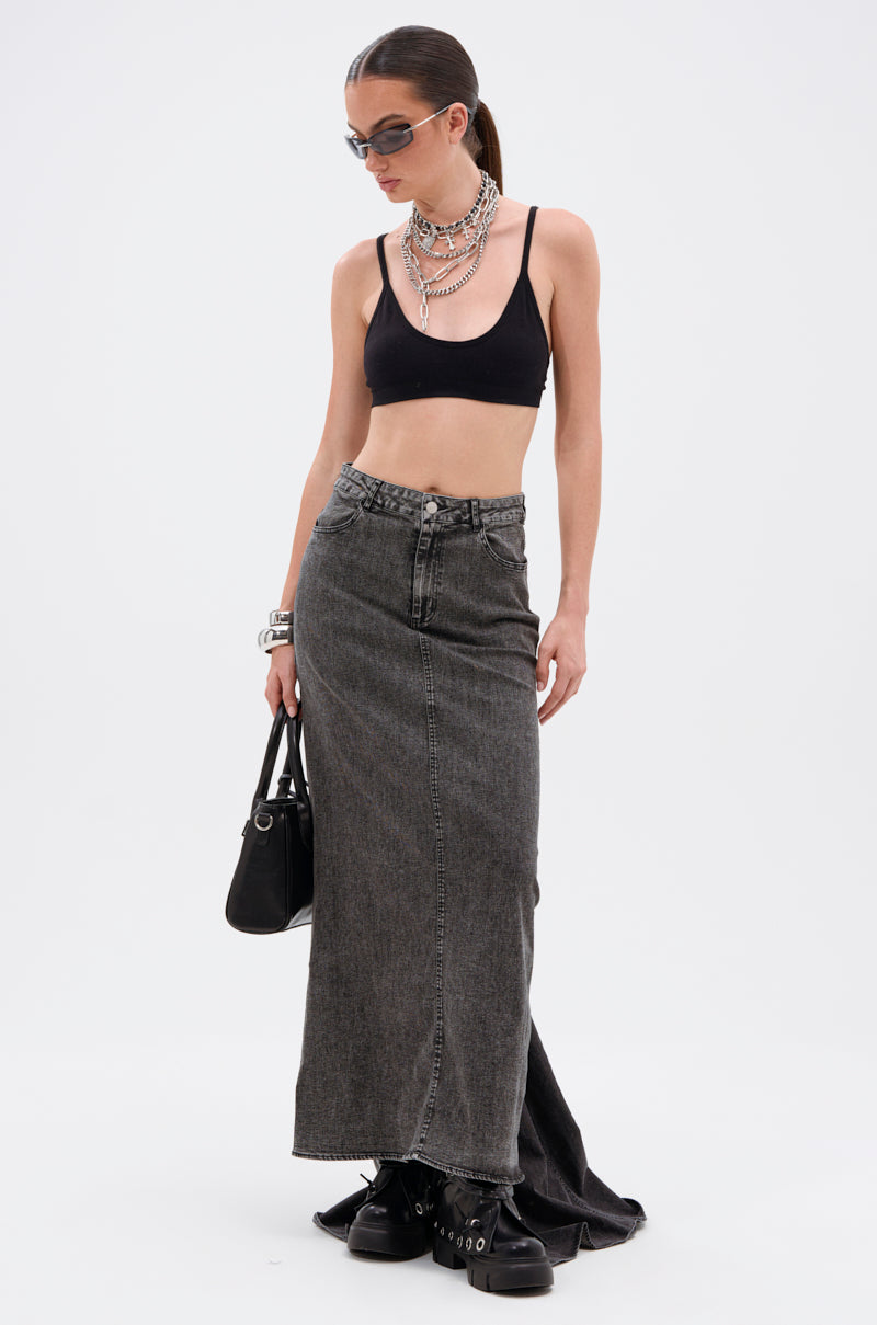 A woman wearing sunglasses, a black crop top, the OH THE DRAMA DENIM MAXI SKIRT WITH TRAIN IN GREY, chunky black boots, layered necklaces and bracelets holds a black handbag while looking down.