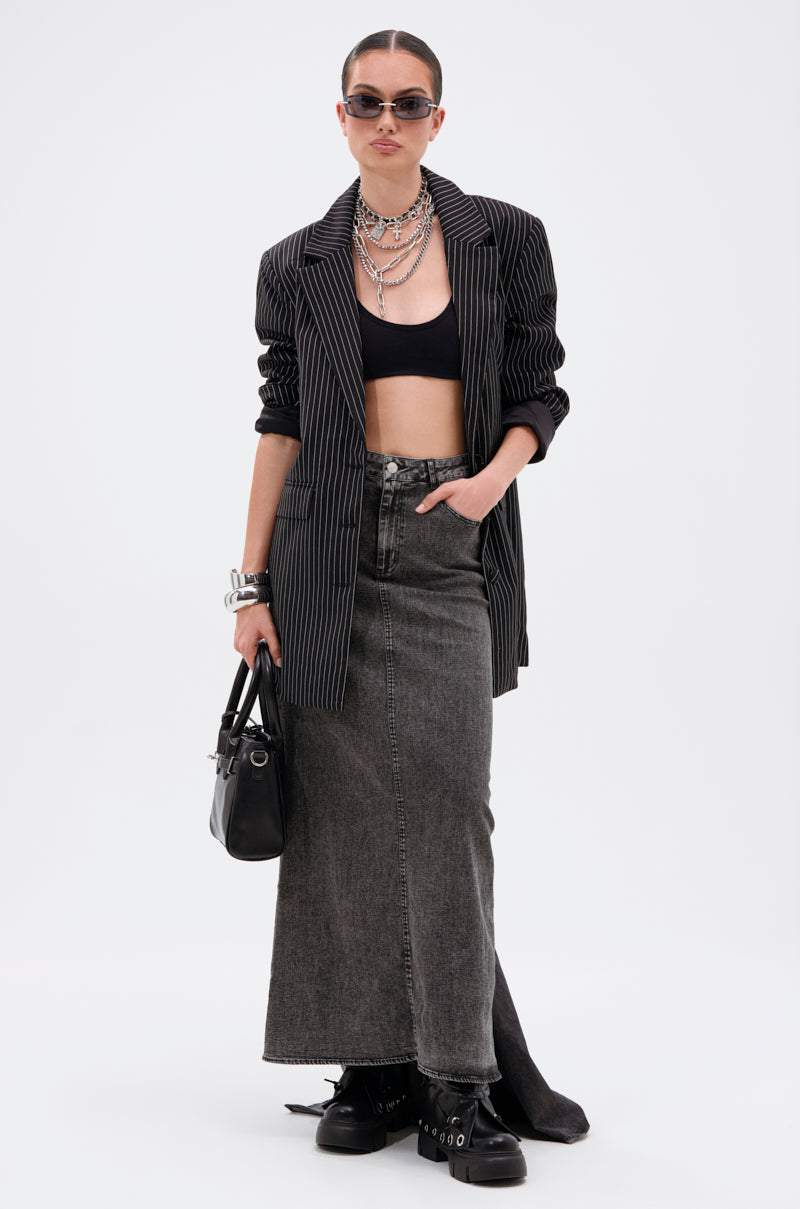 A woman stands against a plain white background wearing sunglasses, a pinstripe blazer over a black crop top, chunky black boots, layered silver jewelry, holding a black handbag, and the OH THE DRAMA DENIM MAXI SKIRT WITH TRAIN IN GREY.