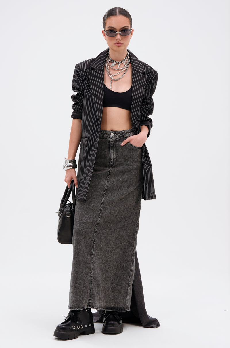 A person wearing sunglasses, layered necklaces, a black crop top, oversized pinstripe blazer, chunky black shoes, silver bracelets, and holding a black handbag stands against a white background in the OH THE DRAMA DENIM MAXI SKIRT WITH TRAIN IN GREY.