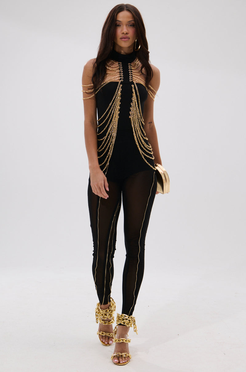 A woman walks forward in the TOO HOT FOR YOU MESH LEGGING, featuring sheer mesh side panels and gold chain embellishments. She carries a gold clutch and wears gold high-heeled sandals with matching chain details against a plain white background. 5