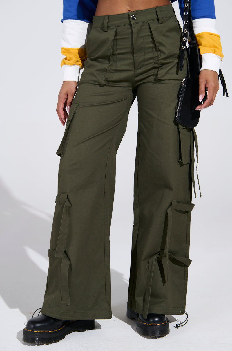 A person wearing IT IS BACK CARGO PANTS in olive green, featuring a high-rise waist, wide legs, and ribbon details, paired with a colorful long-sleeve top. Black platform boots and a matching handbag complete the ensemble.