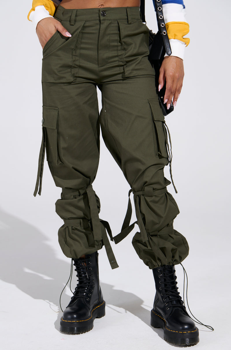 Someone wearing IT IS BACK CARGO PANTS, featuring olive-green color, adjustable drawstring hems, and a high-rise waist, stands against a plain background. They pair the look with black combat boots, a colorful sweatshirt, and a black purse slung over one shoulder.