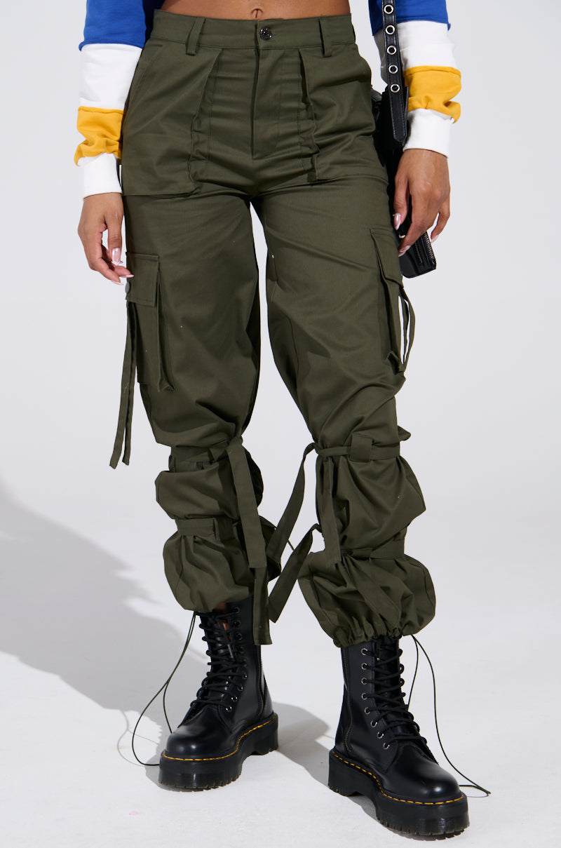 A person wears IT IS BACK CARGO PANTS in olive green with adjustable drawstring hems, black lace-up boots, and carries a sleek black bag. They sport a multicolored long-sleeve top with blue, white, and yellow stripes.