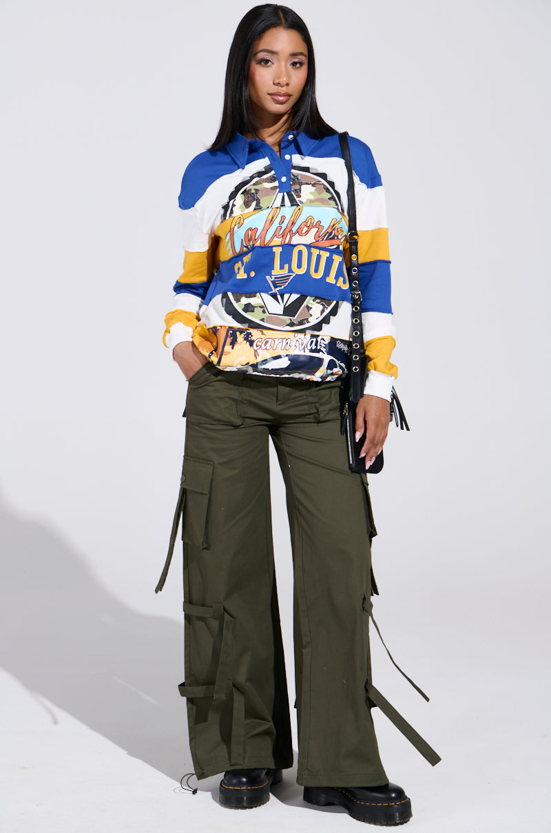 A person in a colorful long-sleeve top blends blue, yellow, and white with IT IS BACK CARGO PANTS in dark green, complete with adjustable drawstring hems. They have long dark hair, hold a black bag, and wear black platform shoes.