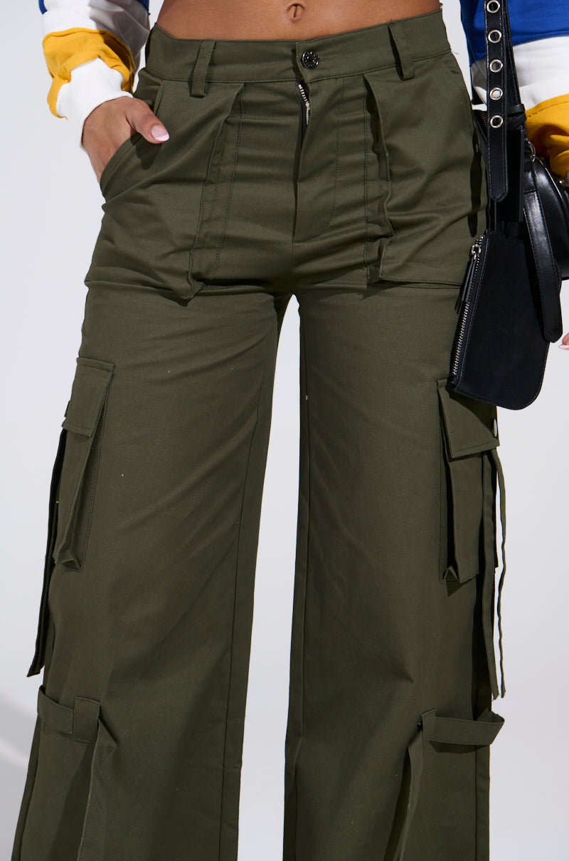 Someone wearing IT IS BACK CARGO PANTS in olive green, featuring multiple pockets and adjustable drawstring hems, paired with a black belt and a large black bag. One hand is tucked into a pocket. The backdrop is a plain white background.