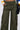 Someone wearing IT IS BACK CARGO PANTS in olive green, featuring multiple pockets and adjustable drawstring hems, paired with a black belt and a large black bag. One hand is tucked into a pocket. The backdrop is a plain white background.