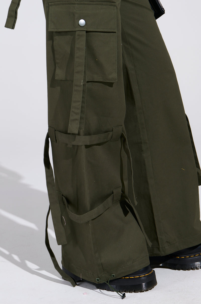 Close-up of a person wearing IT IS BACK CARGO PANTS in olive green, showcasing multiple pockets, straps, and adjustable drawstring hems. Black boots emerge from the bottom against a simple gray backdrop.