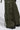 Close-up of a person wearing IT IS BACK CARGO PANTS in olive green, showcasing multiple pockets, straps, and adjustable drawstring hems. Black boots emerge from the bottom against a simple gray backdrop.