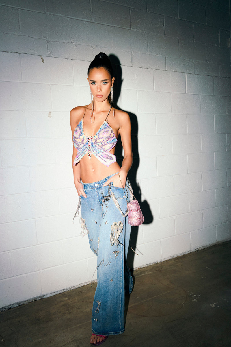 A woman stands against a white brick wall in a butterfly sequin crop top and THE BEST ONE YET RELAXED FIT BLING DENIM JEANS. She holds a sparkly pink purse and gazes confidently at the camera.