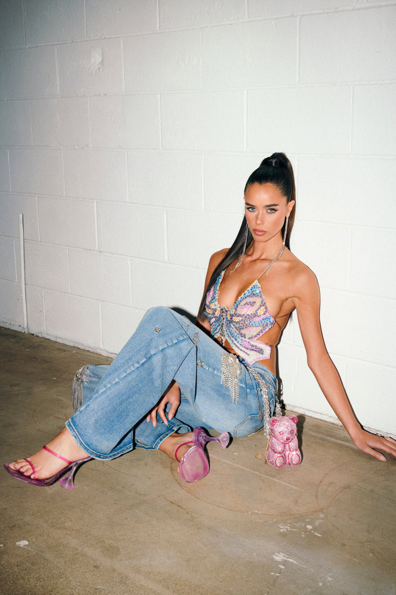 A woman with long dark hair in a high ponytail sits on the floor against a white wall, wearing a colorful butterfly top, pink heels, a small pink bag beside her, and THE BEST ONE YET RELAXED FIT BLING DENIM JEANS.