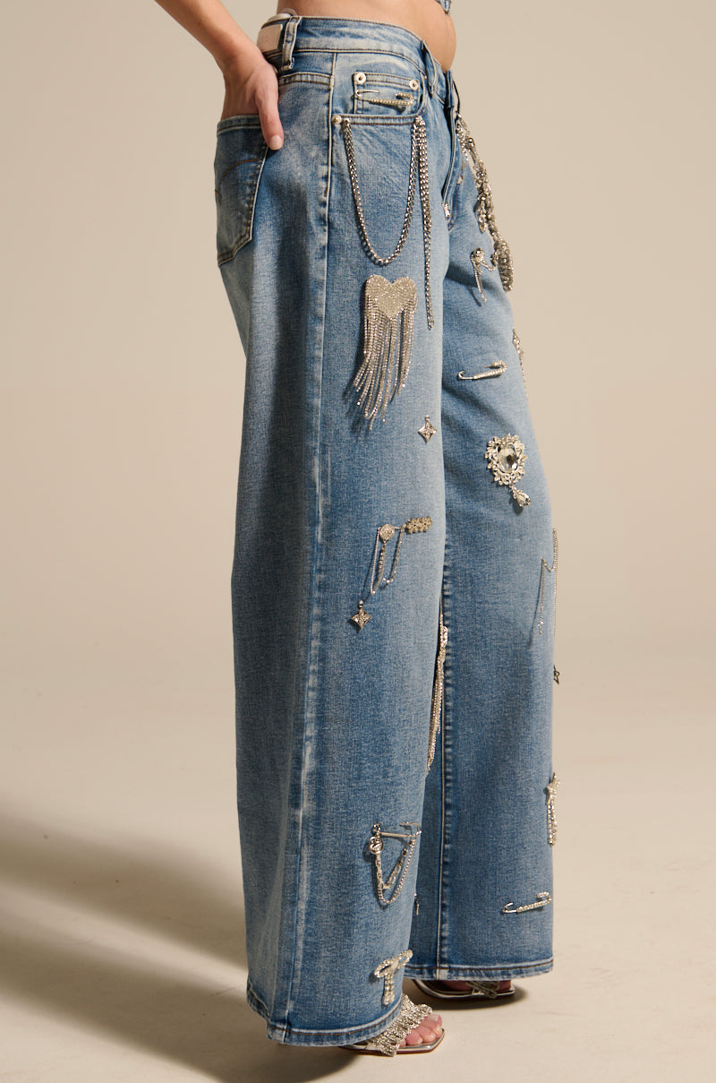Someone wearing THE BEST ONE YET RELAXED FIT BLING DENIM JEANS, featuring metallic chains, wings, and stars embellishments. They stand against a neutral background with one hand in a pocket.
