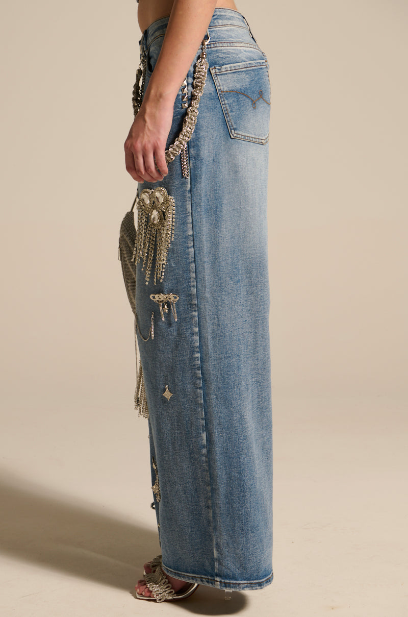 A side view of a long blue denim skirt with silver chains and star embellishments evokes the style of THE BEST ONE YET RELAXED FIT BLING DENIM JEANS. The skirt's back pocket pairs elegantly with open-toe shoes for an effortlessly chic appearance.