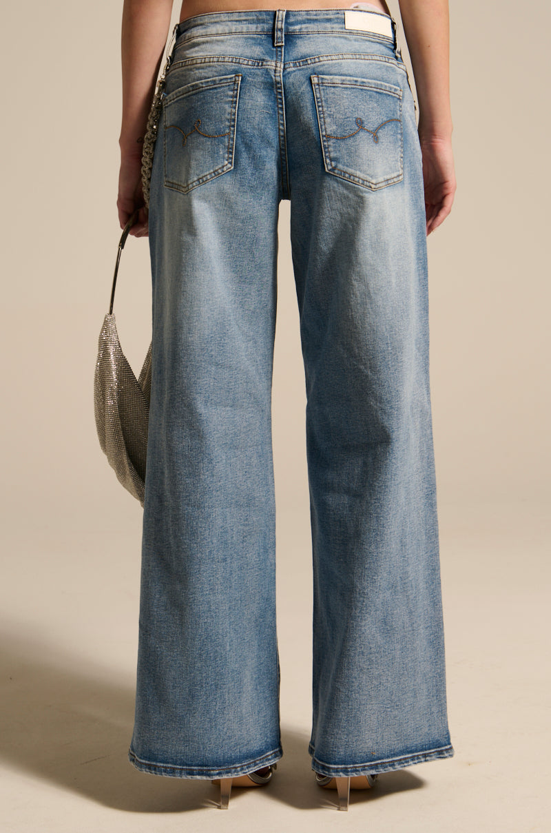 Someone wearing THE BEST ONE YET RELAXED FIT BLING DENIM JEANS with detailed back pockets holds a shimmering silver bag. They stand on a neutral background, beige high heels visible, showcasing their stylish ensemble.