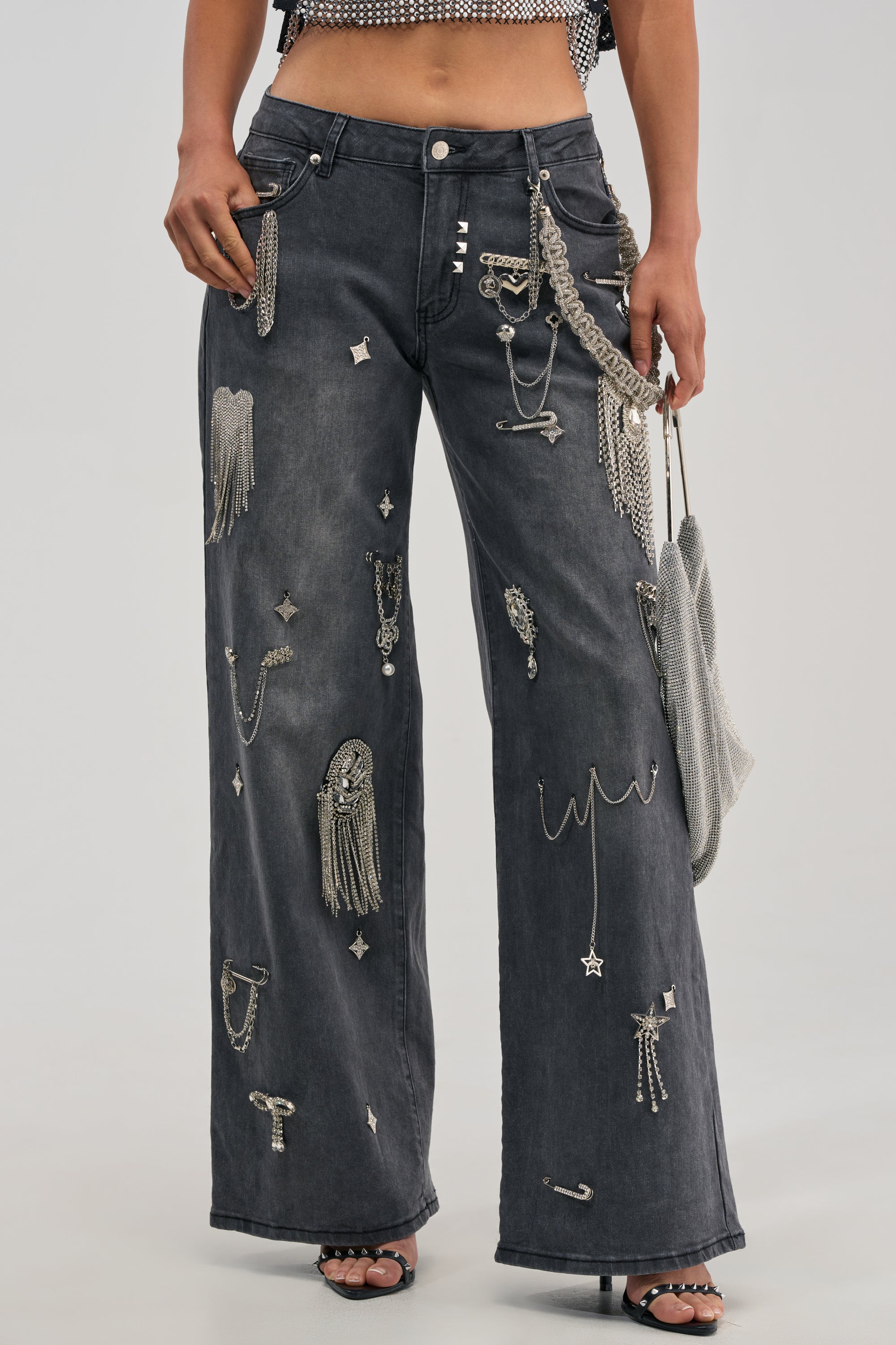A person wearing THE BEST ONE YET RELAXED FIT BLING DENIM JEANS—black wide-leg streetwear adorned with silver chains, studs, and metallic embellishments—holds a chainmail-style handbag. Only the lower torso and legs are visible against a plain background.