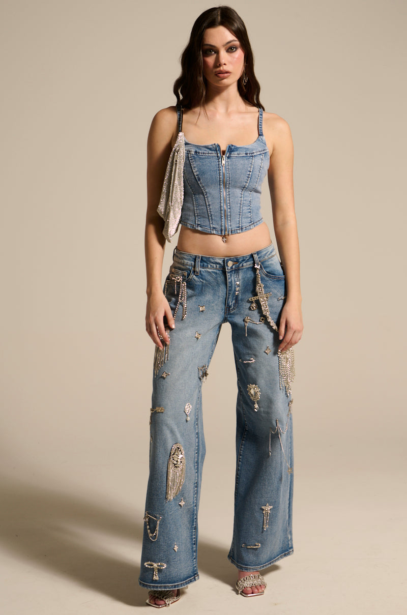 A woman models THE BEST ONE YET DENIM CORSET with a metallic zipper closure and embellished jeans featuring shiny decorations. Her long, wavy hair flows down, set against a plain background, showcasing a fashion-forward Y2K-inspired style.