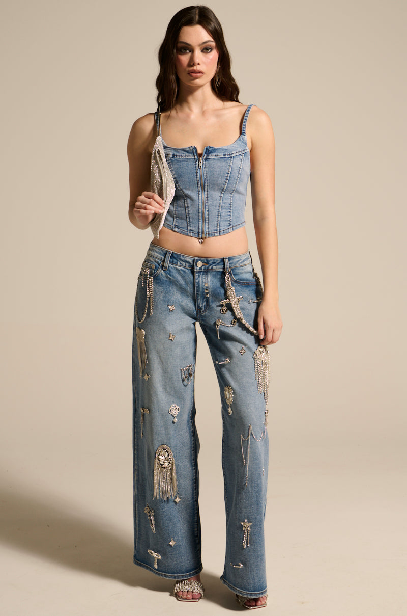 A woman in "THE BEST ONE YET DENIM CORSET" with a metallic zipper closure pairs it with wide-leg jeans adorned with sparkling embellishments. She stands against a plain background, holding a small purse and wearing open-toe heels. Her loose hair and confident expression echo the Y2K style.