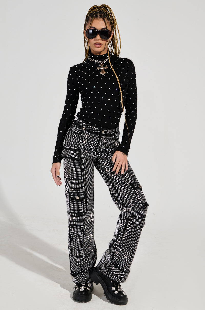A person confidently poses in a black, star-patterned long-sleeve top paired with black Ammo Bonne Soiree Rhinestone Cargo Pants, featuring a high-rise waist. They showcase chunky shoes, long braided hair, and sunglasses against a plain, light-colored backdrop.