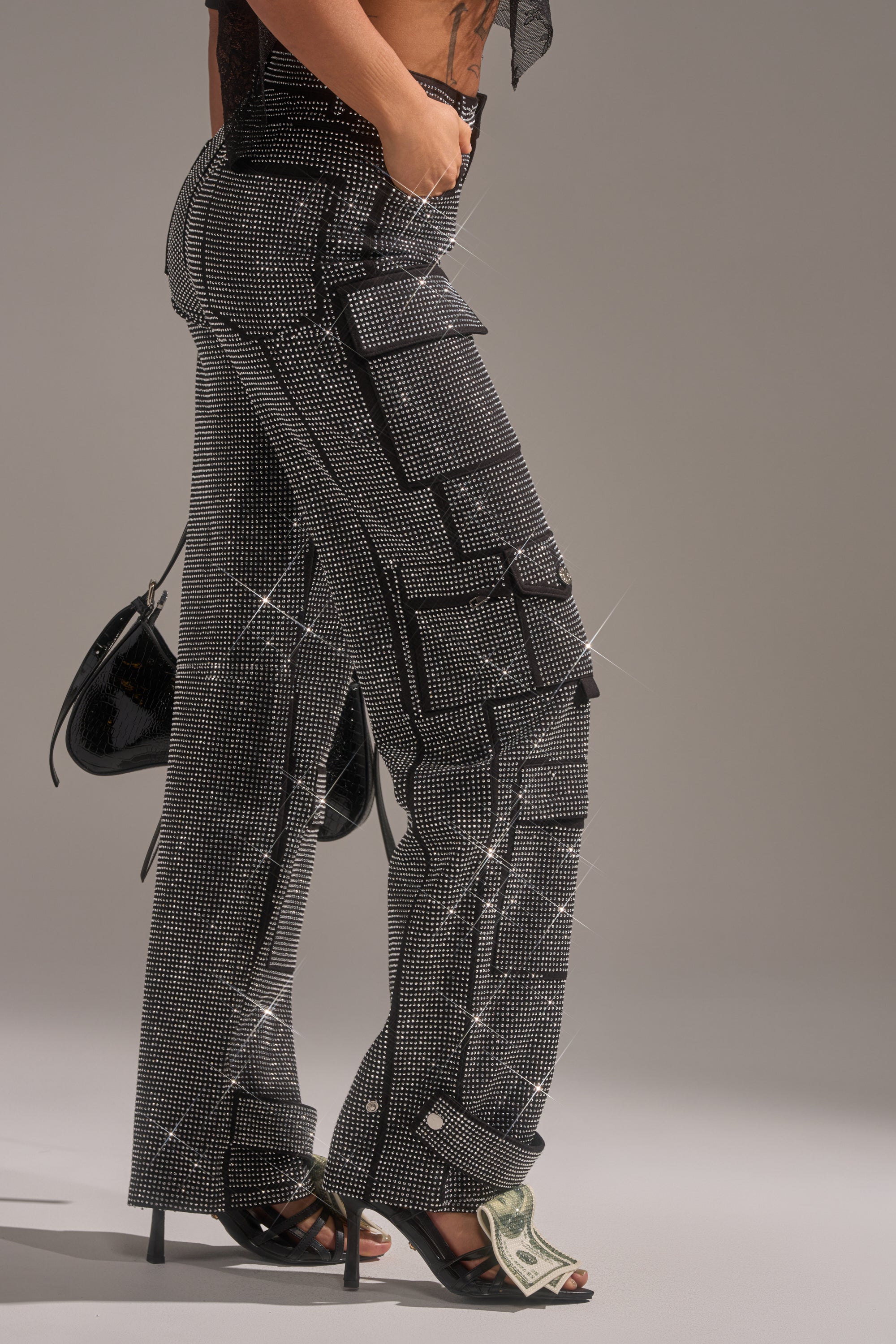 Wearing AMMO BONNE SOIREE RHINESTONE CARGO PANTS IN BLACK, a person pairs them with black heeled sandals—dollar bill underfoot—and holds a small black purse, all set against a plain background.