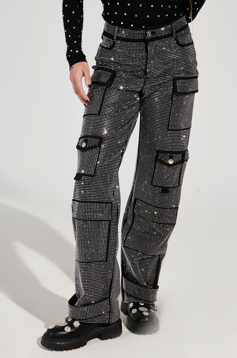 The individual is wearing the AMMO BONNE SOIREE Rhinestone Cargo Pants in Black, featuring a high-rise waist and multiple pocket designs with glossy buttons. The look is paired with a black, long-sleeved polka dot shirt and black shoes adorned with decorative elements.
