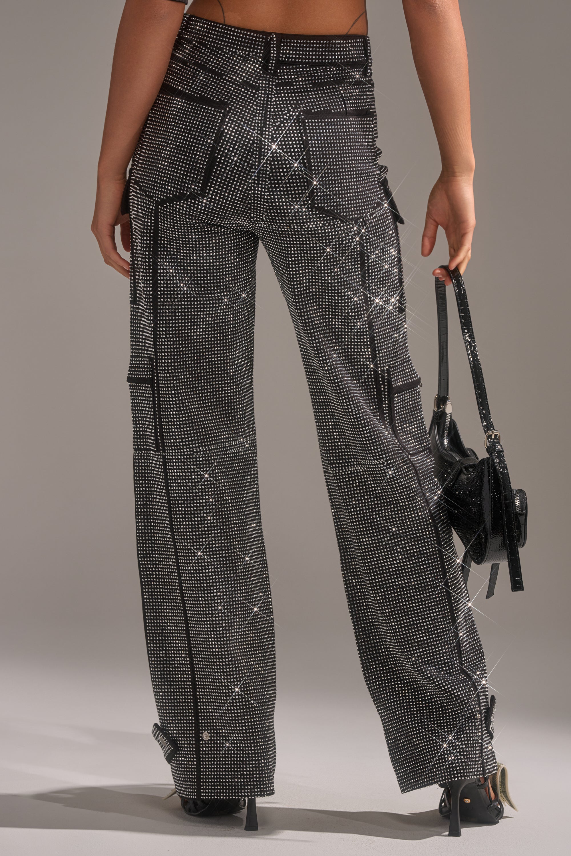 A person faces away in AMMO BONNE SOIREE RHINESTONE CARGO PANTS IN BLACK, styled with a small black bag and black heels against a plain, neutral background.