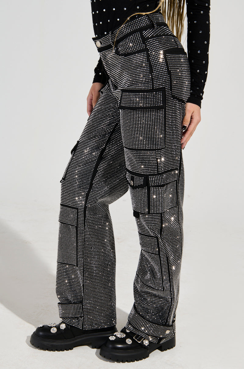 The individual is dressed in the AMMO BONNE SOIREE RHINESTONE CARGO PANTS IN BLACK, featuring a grid pattern adorned with sparkly rhinestones, paired with a black long-sleeve shirt decorated with white polka dots. Their ensemble is completed with black boots embellished with silver accents, all set against a simple, light-colored background.