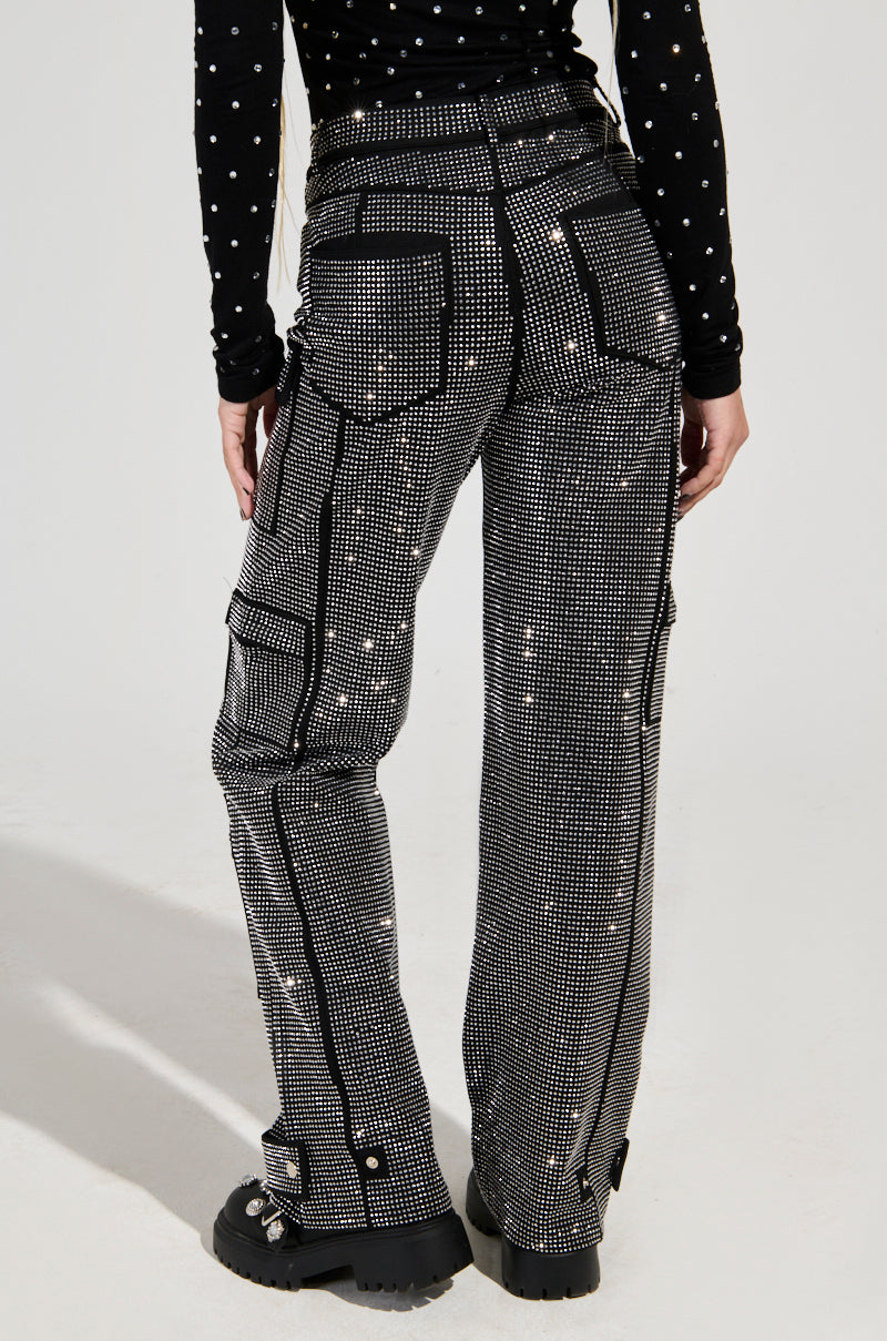 A person is sporting the AMMO BONNE SOIREE RHINESTONE CARGO PANTS IN BLACK, which feature white polka dots, studded details, and a high-rise waist with visible pockets and zippers. They’ve coordinated it with a matching long-sleeve shirt and finished the look with chunky black boots.