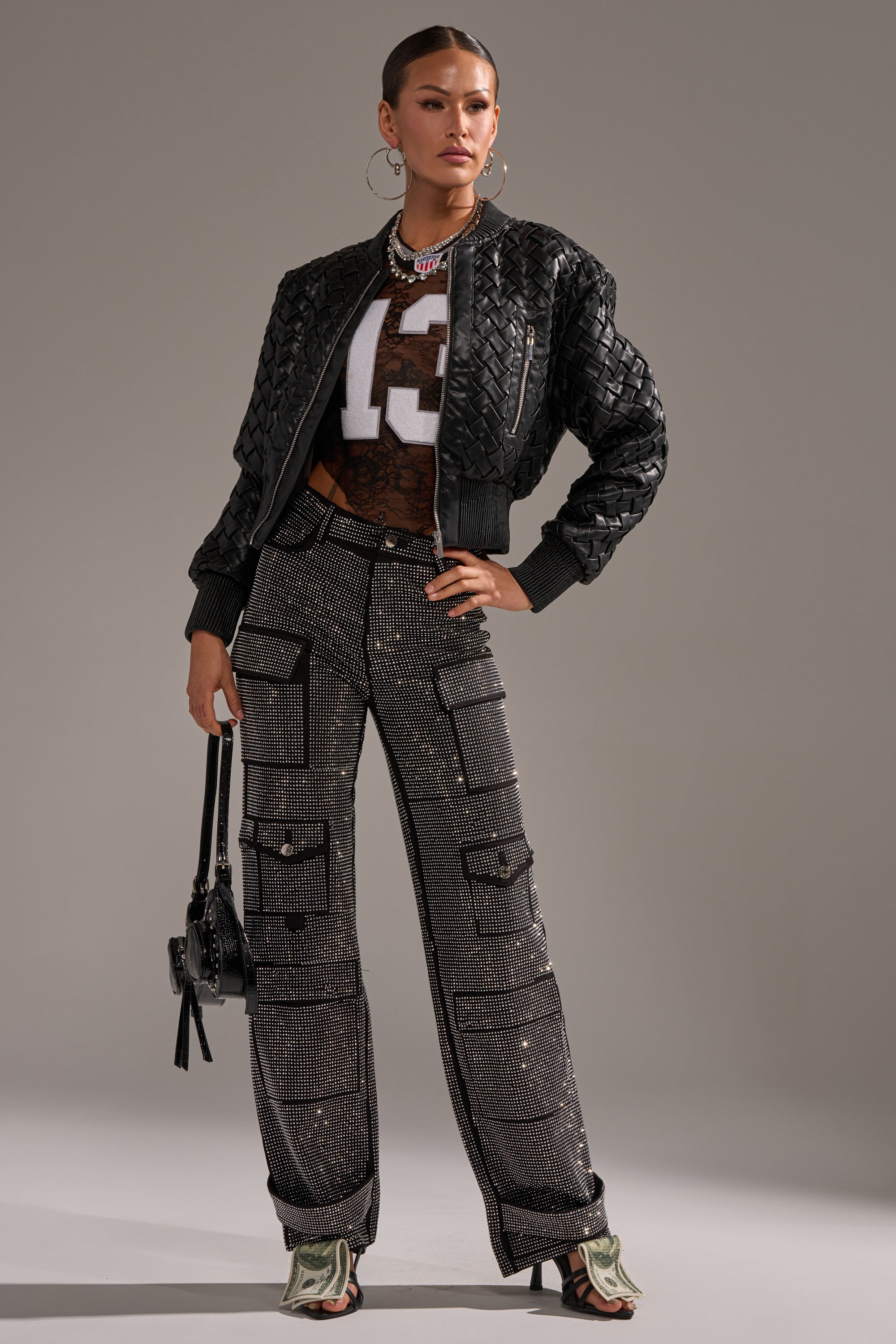 A woman poses in AMMO BONNE SOIREE RHINESTONE CARGO PANTS IN BLACK, a black textured jacket, sheer top with "13", hoop earrings, a small handbag, and open-toe heeled boots against a plain background.