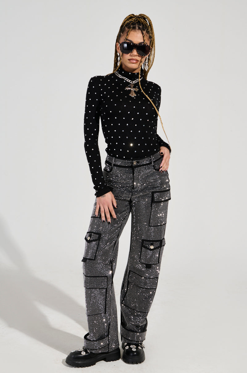 A person with long braided hair wears black sunglasses, a black long-sleeve top adorned with white polka dots, and the AMMO BONNE SOIREE RHINESTONE CARGO PANTS IN BLACK, known for their high-rise waist. They stand confidently against a plain white background.