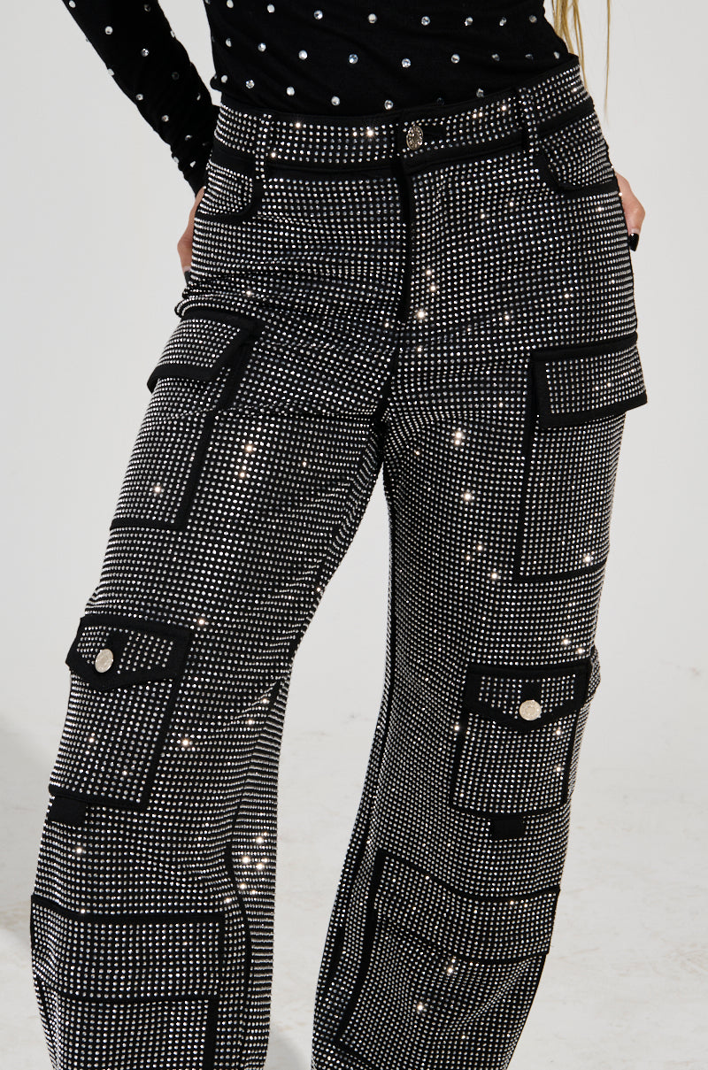 In the image, an individual is wearing a black and white polka dot top paired with the AMMO BONNE SOIREE Rhinestone Cargo Pants in Black. These high-rise waist pants feature a sparkling design with shimmering, reflective dots and multiple pockets. The stylish outfit is made from stretch knit material, offering an eye-catching look.