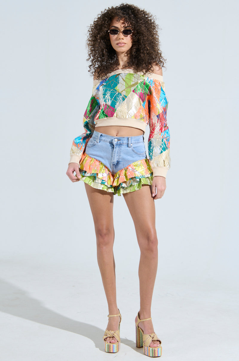 Someone with curly hair sports sunglasses, a colorful off-the-shoulder sweater, LIFE IS BUT A DREAM BROCADE RUFFLE DENIM SHORTS, and platform sandals. They stand confidently against a plain background.