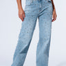 A person is modeling the EVERYTIME RHINESTONE EMBELLISHED STRAIGHT LEG JEANS in a high rise light blue wash, adorned with numerous small studs. They are also wearing a black leather vest and black leather shoes with studded details. The image is cropped at the waist and feet, focusing on the jeans.