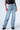 The individual is dressed in the EVERYTIME RHINESTONE EMBELLISHED STRAIGHT LEG JEANS, characterized by their light blue color and numerous silver studs. They also have on a black leather vest and black chunky shoes with silver embellishments, standing against a plain white background.