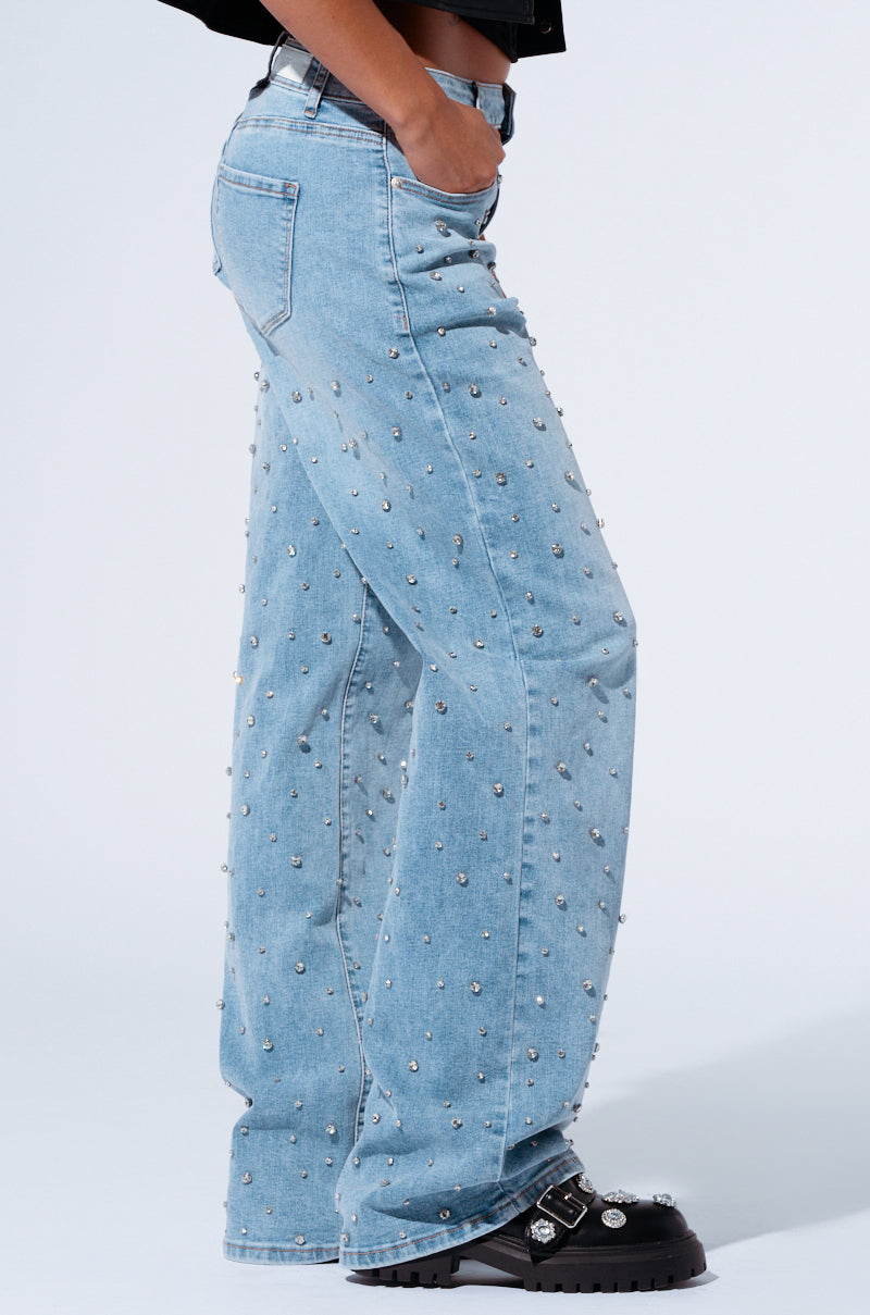 A person is standing in the EVERYTIME RHINESTONE EMBELLISHED STRAIGHT LEG JEANS, which are light blue and decorated with small round studs. With one hand in their pocket, they are also wearing black leather shoes featuring multiple buckles. The background is plain white.