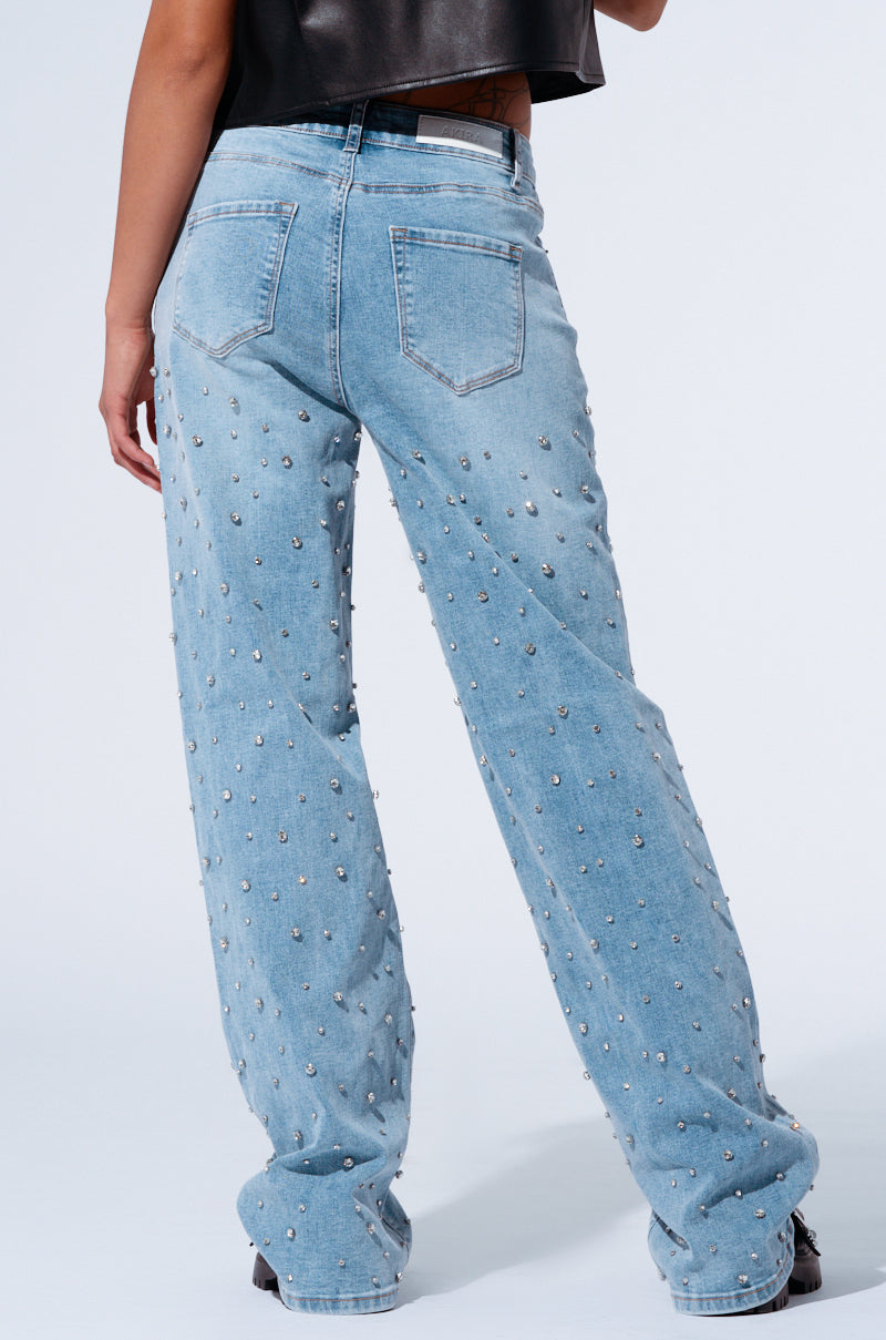 A person is shown from the waist down, wearing EVERYTIME RHINESTONE EMBELLISHED STRAIGHT LEG JEANS in light blue, featuring metallic studs. They are also wearing a black top. The loose-fitting jeans have visible back pockets and the person is standing against a plain, light background.