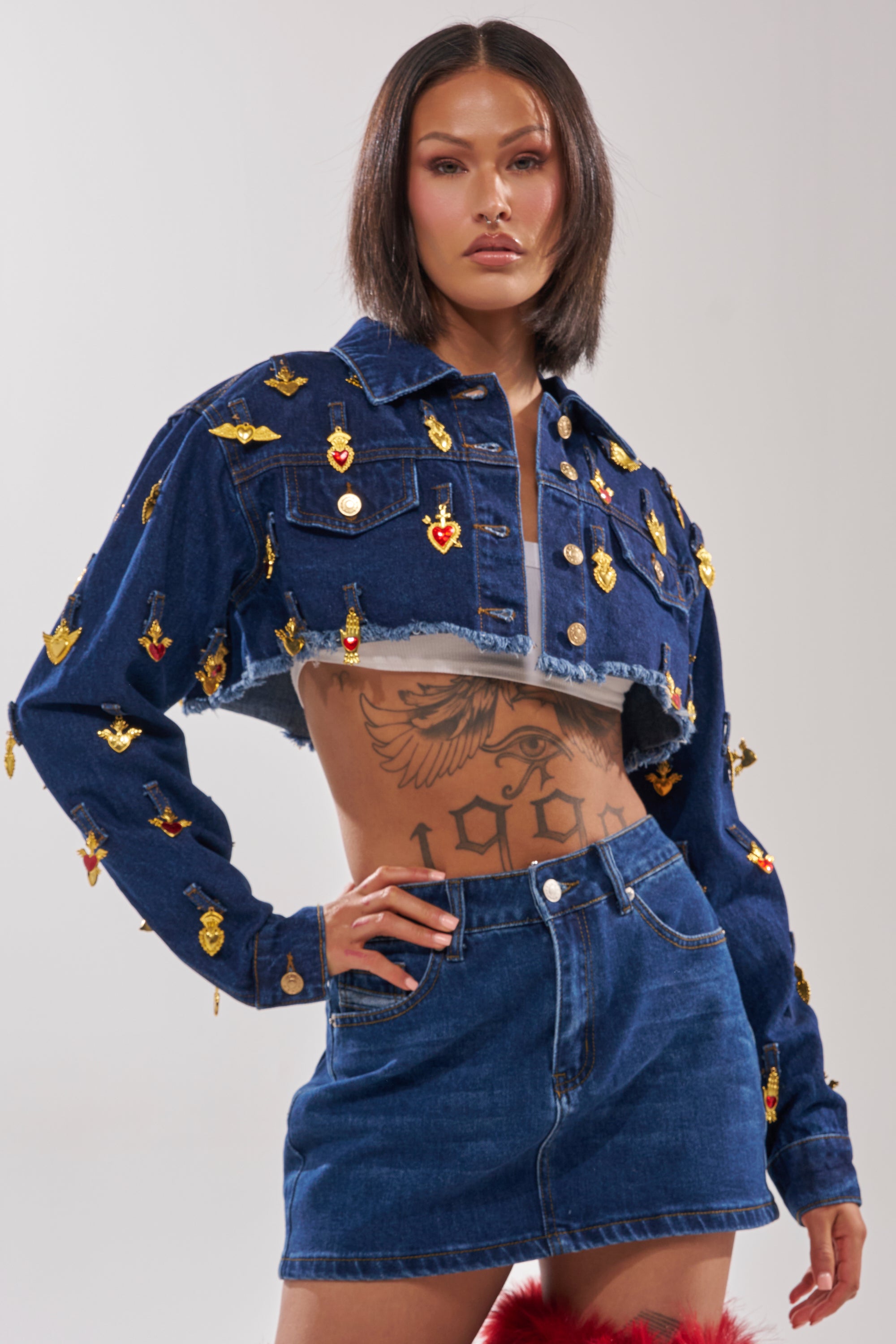 A person with straight dark hair wears the JUST A CRUSH CROPPED DENIM JACKET with gold pins and a matching skirt. Tattoos adorn their torso as they stand confidently, one hand on their hip. 0