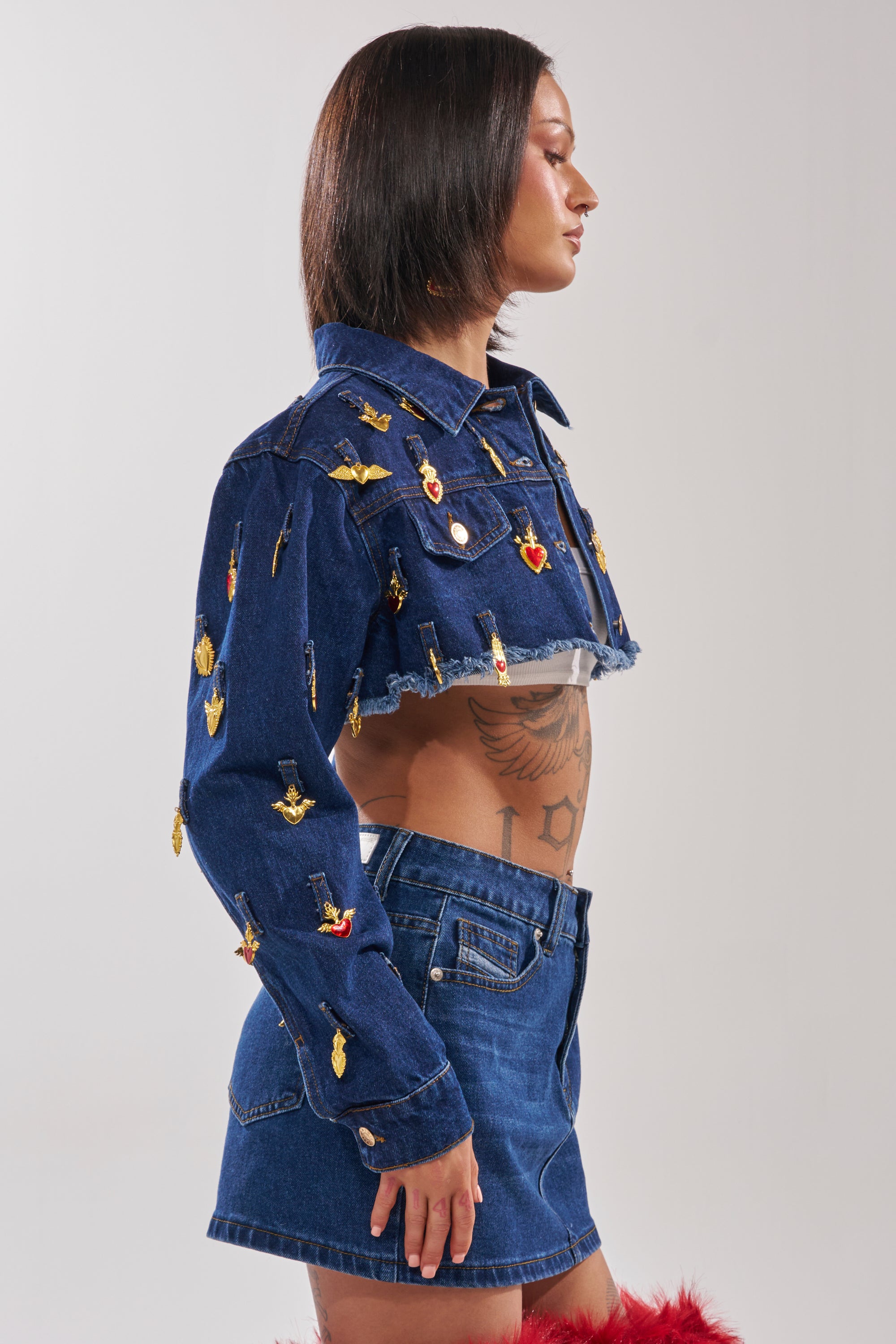 A woman with straight dark hair poses in profile against a light background, wearing the JUST A CRUSH CROPPED DENIM JACKET adorned with pins and a matching skirt, revealing her torso tattoos. 2