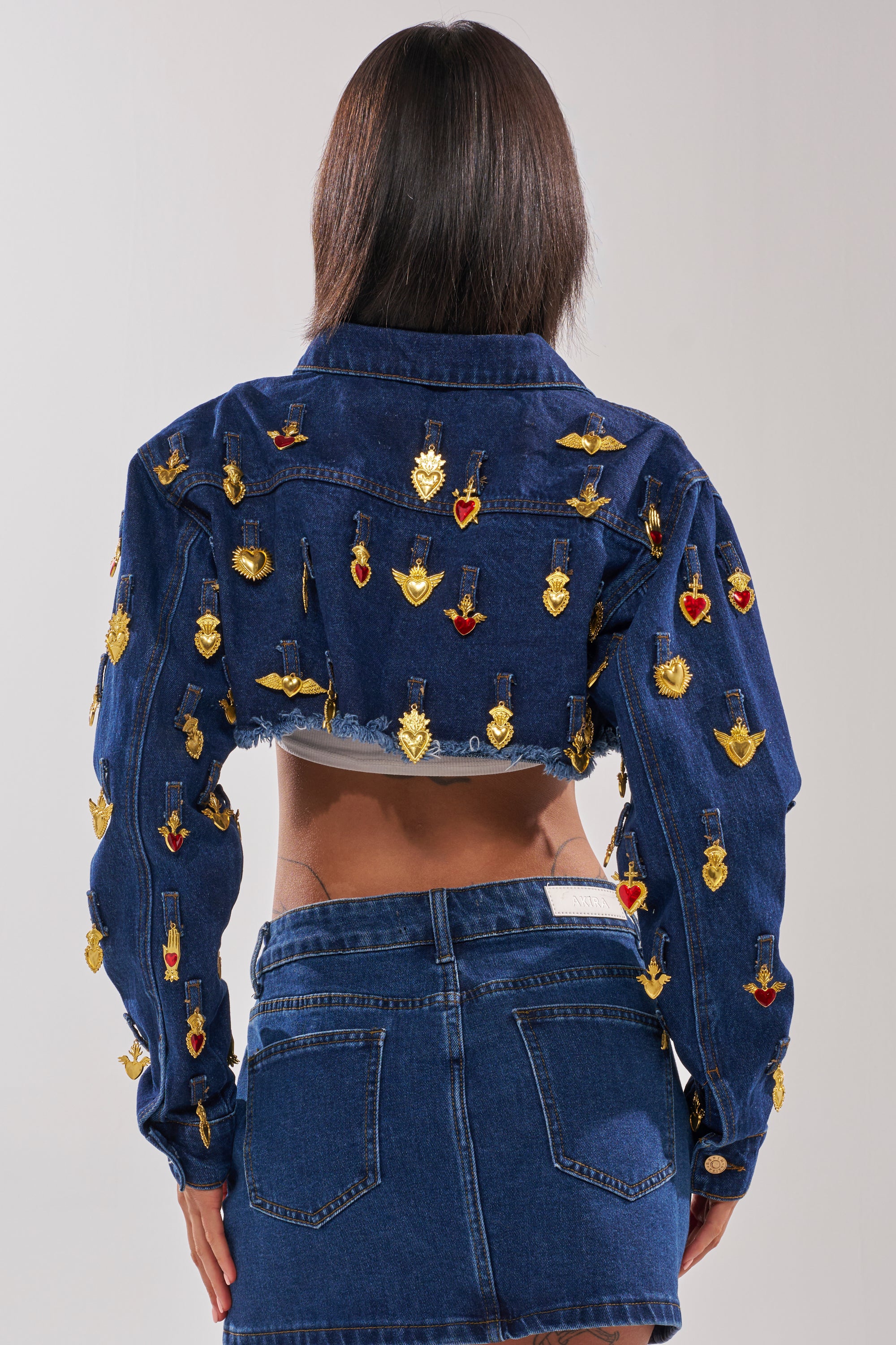A woman faces away from the camera, wearing the JUST A CRUSH CROPPED DENIM JACKET, which features bold gold and red heart-shaped embellishments. 3