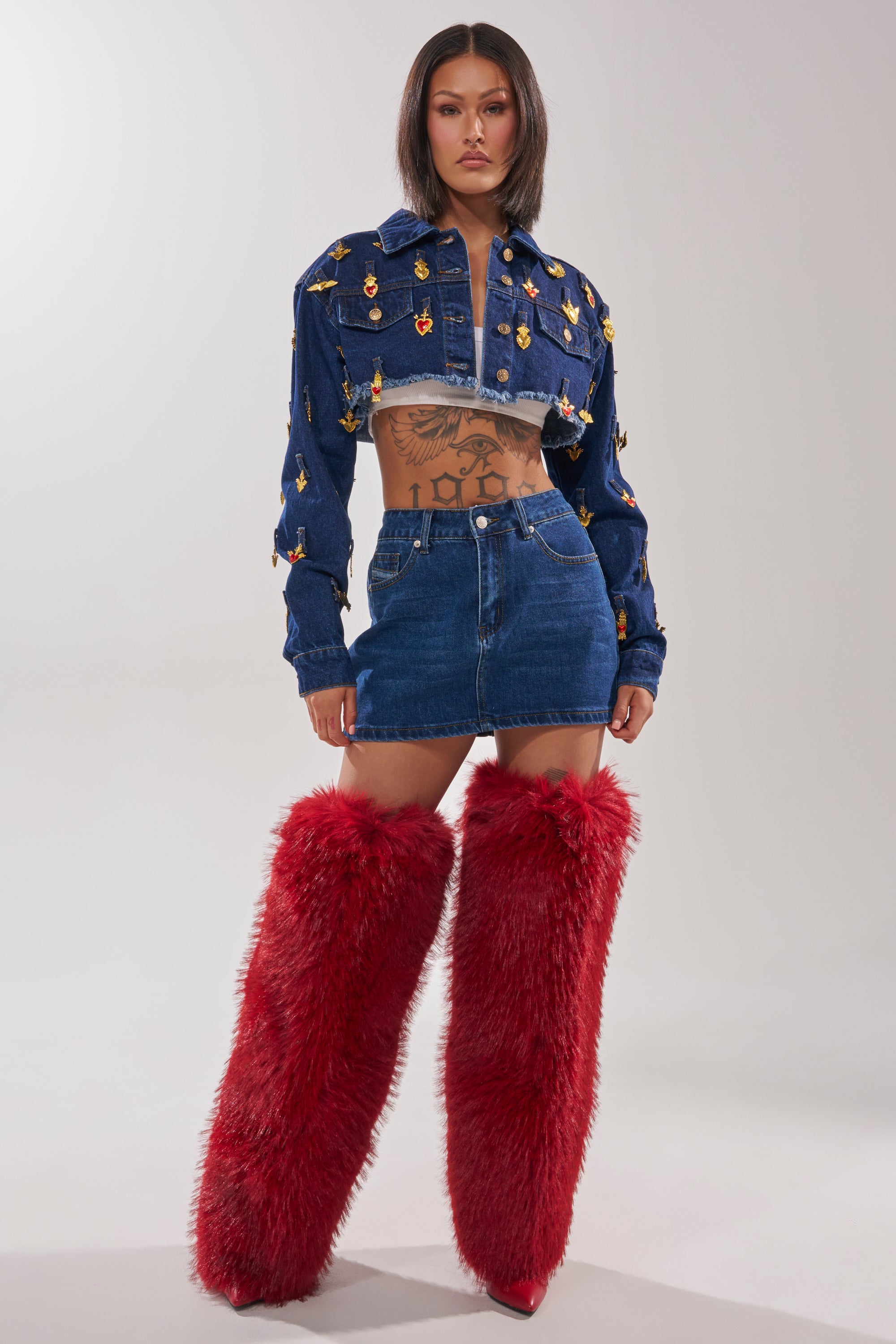 A woman models the JUST A CRUSH CROPPED DENIM JACKET with a coordinating mini skirt and tall red fur boots, showcasing her straight dark hair and midriff tattoos against a simple light-colored backdrop. 4