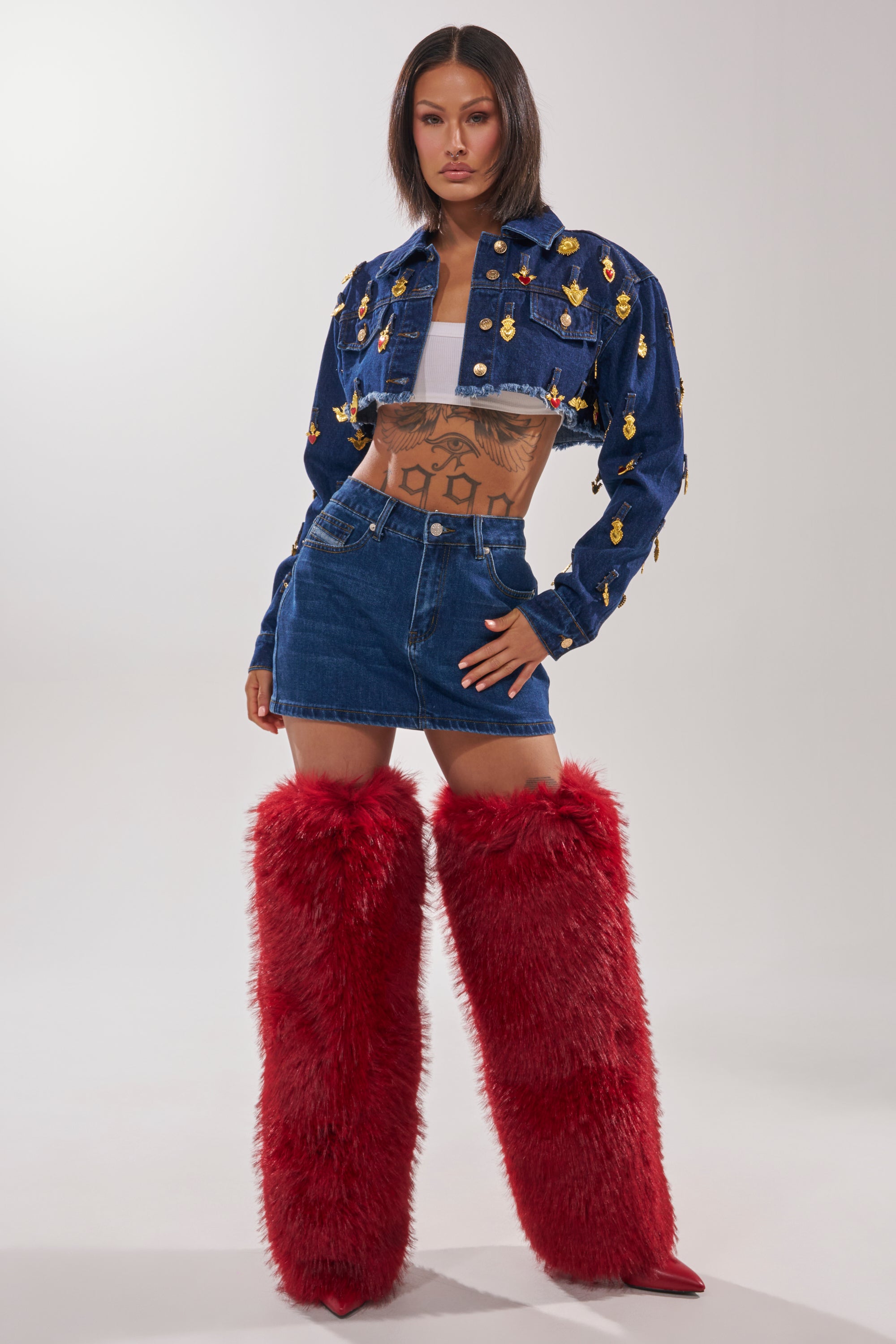 A woman stands with hands on hips, showing tattoos and wearing the JUST A CRUSH CROPPED DENIM JACKET with gold embellishments, a white crop top, denim mini skirt, and tall red furry boots. 5