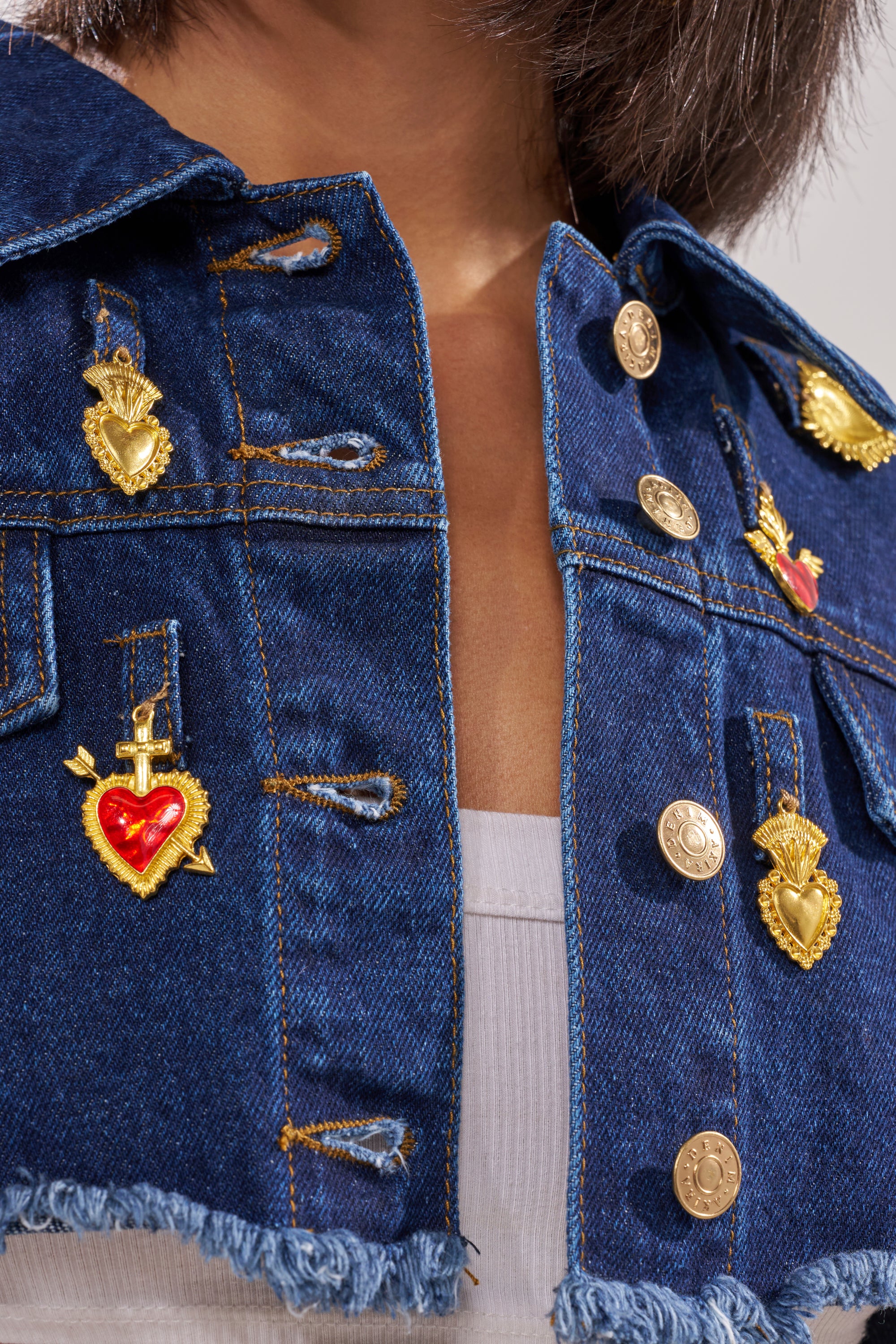 A close-up of a person wearing the JUST A CRUSH CROPPED DENIM JACKET, embellished with gold and red heart-shaped pins, over a white ribbed top. Only the lower face and neck are visible. 6