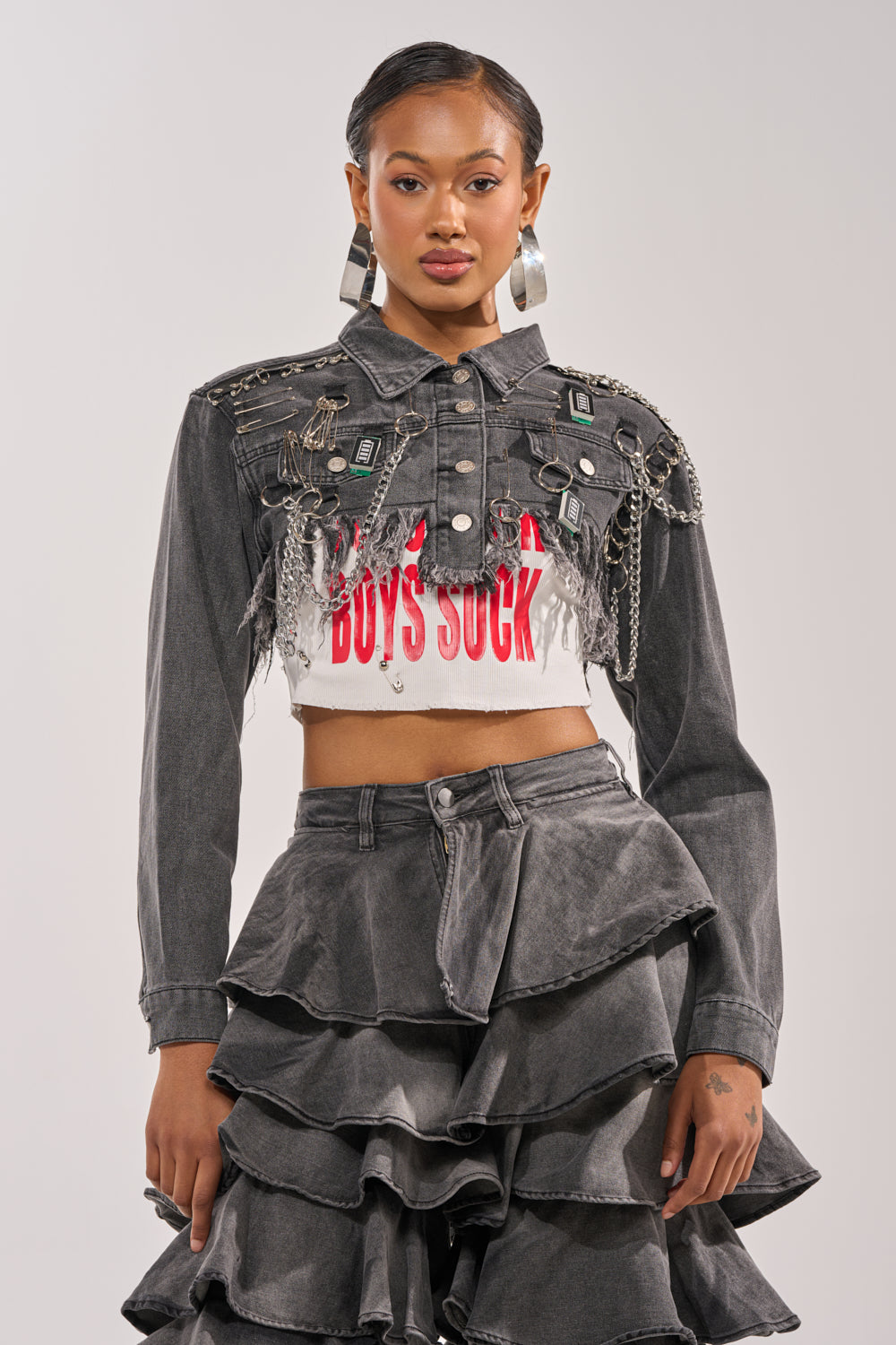 A woman wears the CAN'T COMPUTE ULTRA CROPPED DENIM JACKET with a distressed hemline over a white "BOYS SUCK" crop top, styled with a tiered black ruffled skirt and large hoop earrings. 0