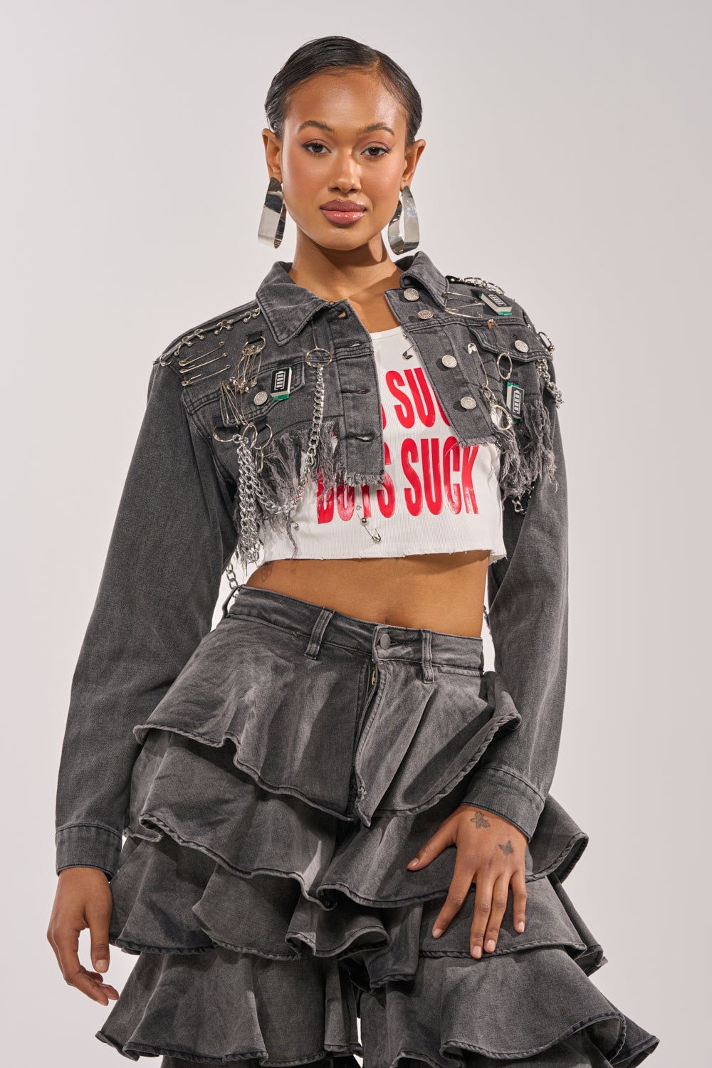 A woman poses confidently in the CAN'T COMPUTE ULTRA CROPPED DENIM JACKET with embellishments and a distressed hem, a white "BOYS SUCK" crop top, and a gray tiered ruffled denim skirt. 1