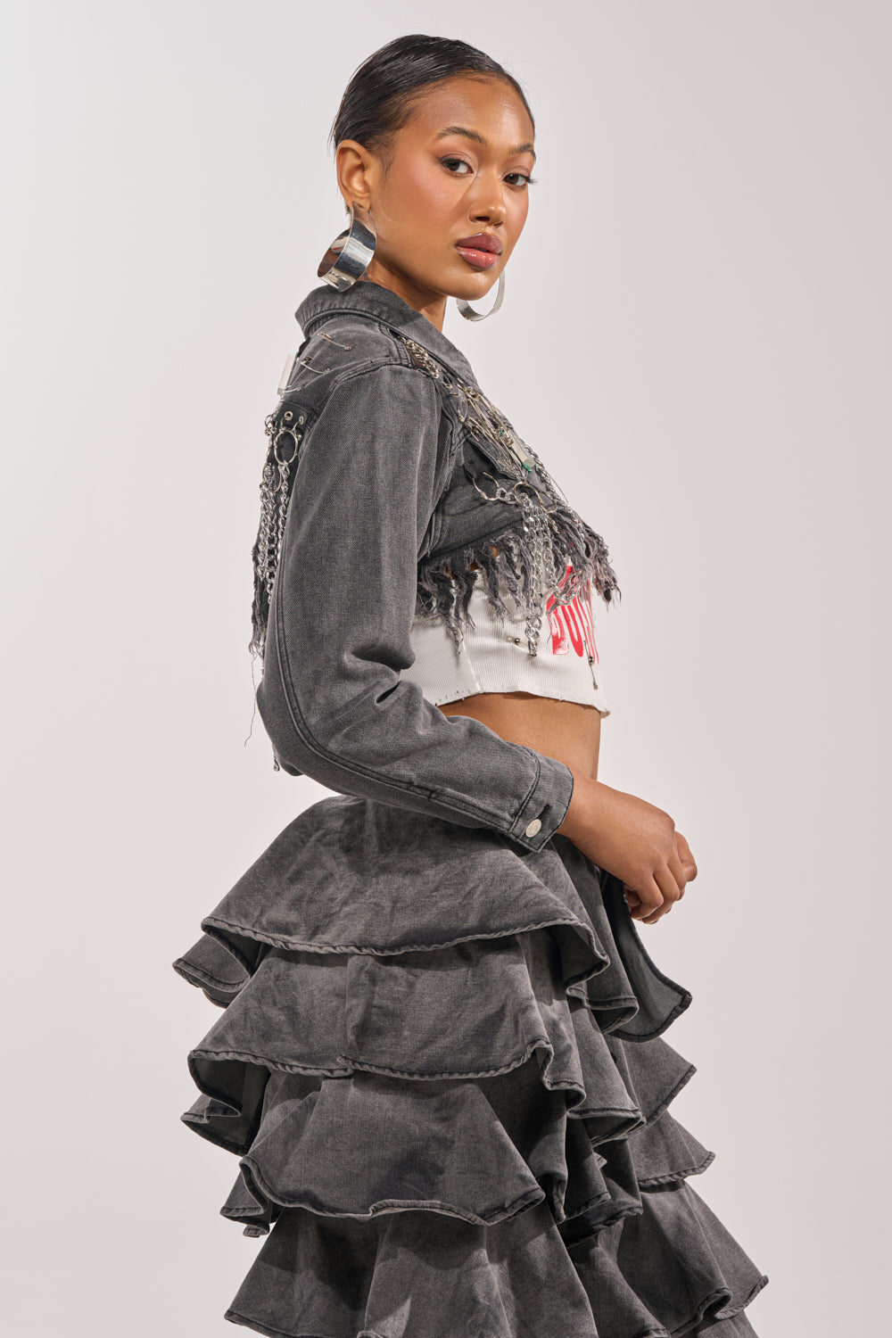 A woman poses in side profile wearing the CAN'T COMPUTE ULTRA CROPPED DENIM JACKET with chain details, paired with a white crop top and high-waisted gray ruffle skirt. She accessorizes with hoop earrings and a sleek hairstyle. 2