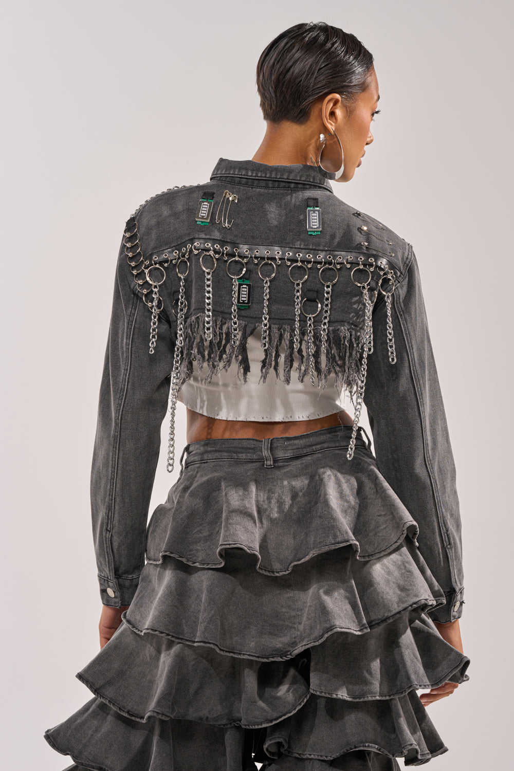 A person with neatly styled short hair faces away, wearing the CAN'T COMPUTE ULTRA CROPPED DENIM JACKET embellished with studs, chains, and electronic components, paired with a tiered ruffle skirt featuring a distressed hemline. 3