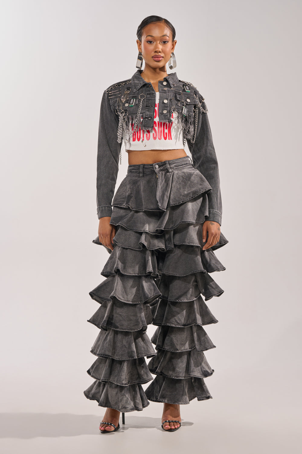 A woman wears the CAN'T COMPUTE ULTRA CROPPED DENIM JACKET with fringe and a distressed hem, a white crop top with red text, high-waisted gray ruffled pants layered in tiers, and open-toe heels. 4